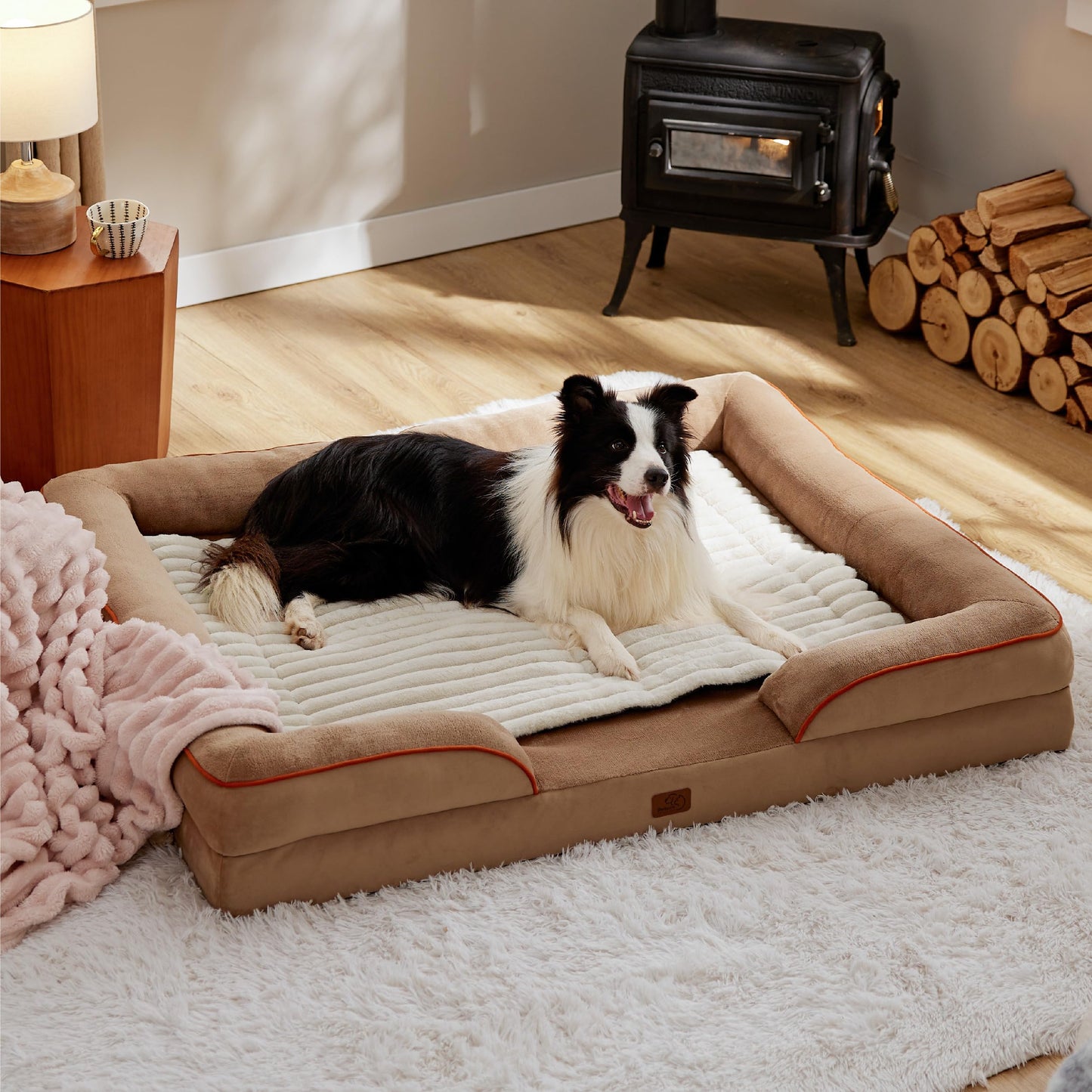 Dog lying on a large beige pet bed in a cozy room with a fireplace and wooden logs.